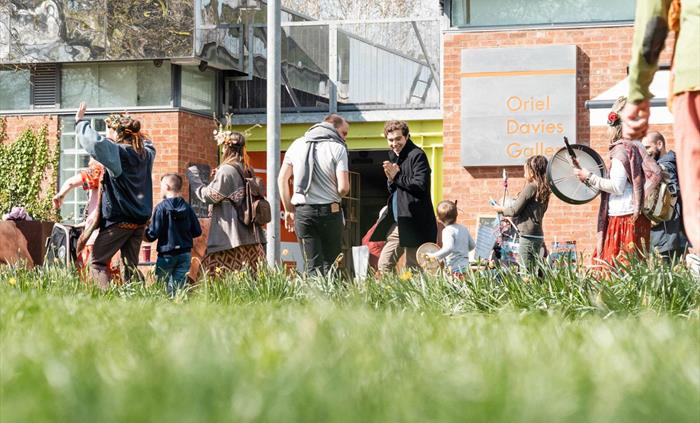 Looking through grass in the foreground Floor level view of people walking in procession in front of the gallery wearing flower crowns carrying and pl